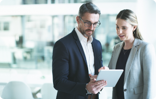 Two business professionals, a man and a woman, collaborate on a tablet in a modern office setting.