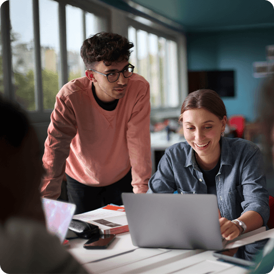 Two people working together at a laptop in a dimly lit room with large windows.